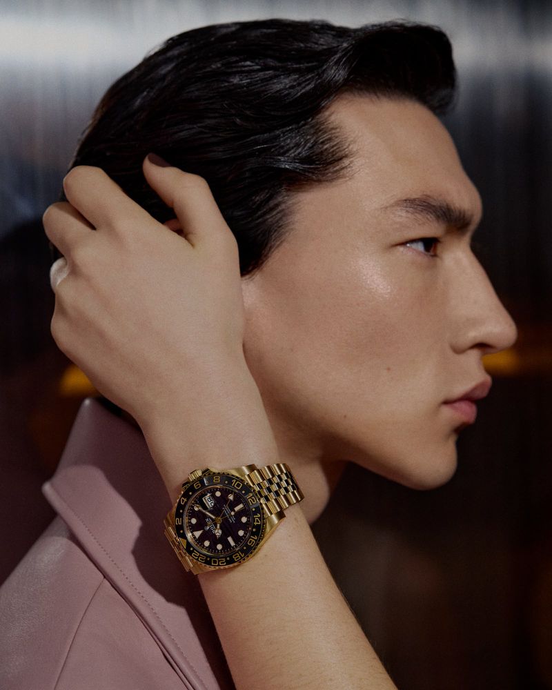 Close-up of an Asian male model wearing a pink jacket and a gold Rolex watch, hand on his head, shot in a photo studio.
