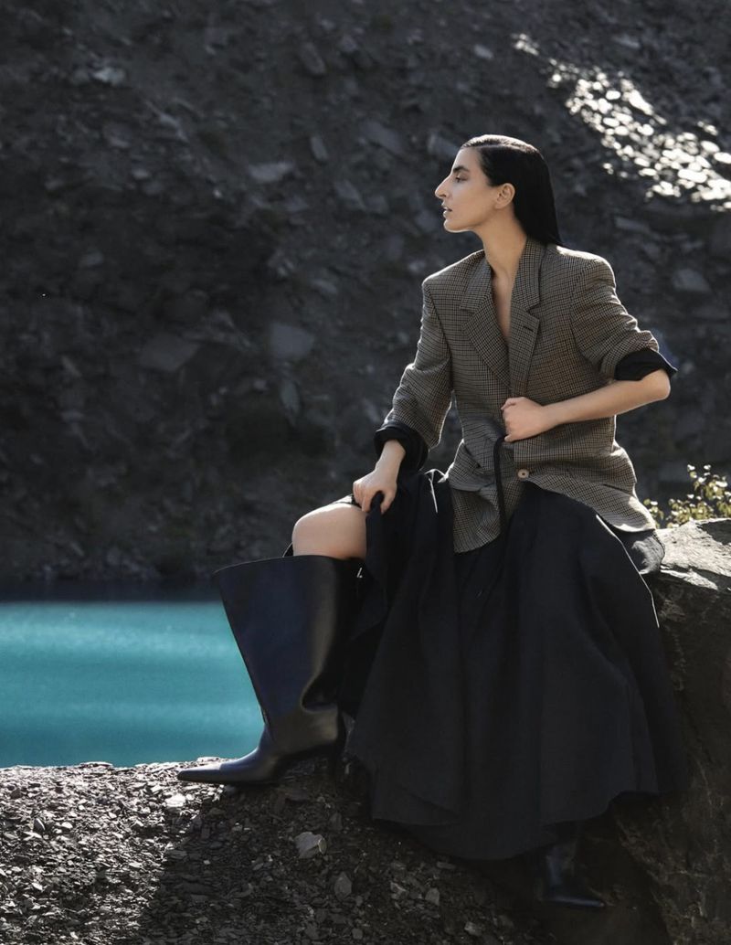 Woman in blazer and skirt posing on rocky terrain near aqua blue water with dark, rocky hillside in background in a fashion editorial photo.