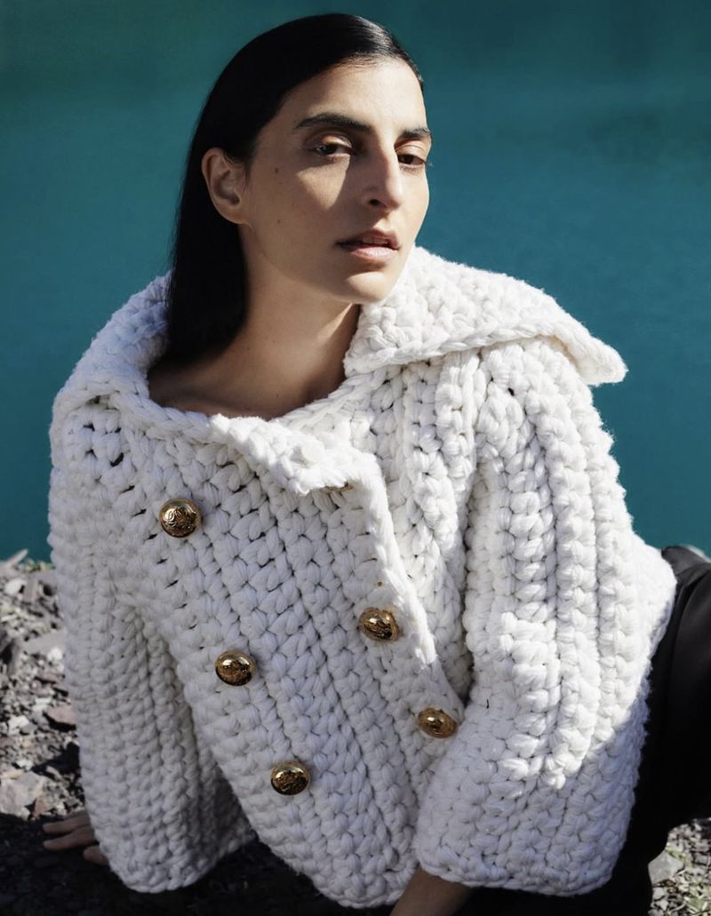 A woman poses outdoors wearing a white, double-breasted knit jacket with gold buttons, against a blue-green water background.
