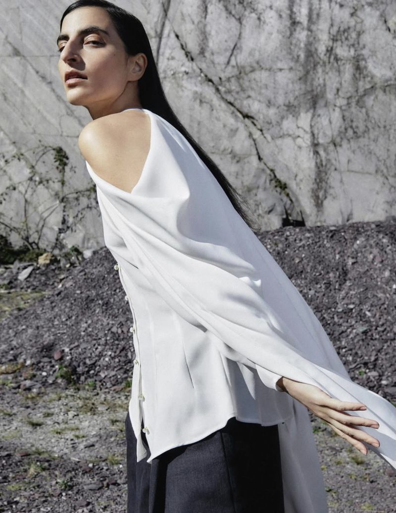 A young adult female model poses in an off-shoulder white top and dark pants outdoors, set against a striking rock wall backdrop.