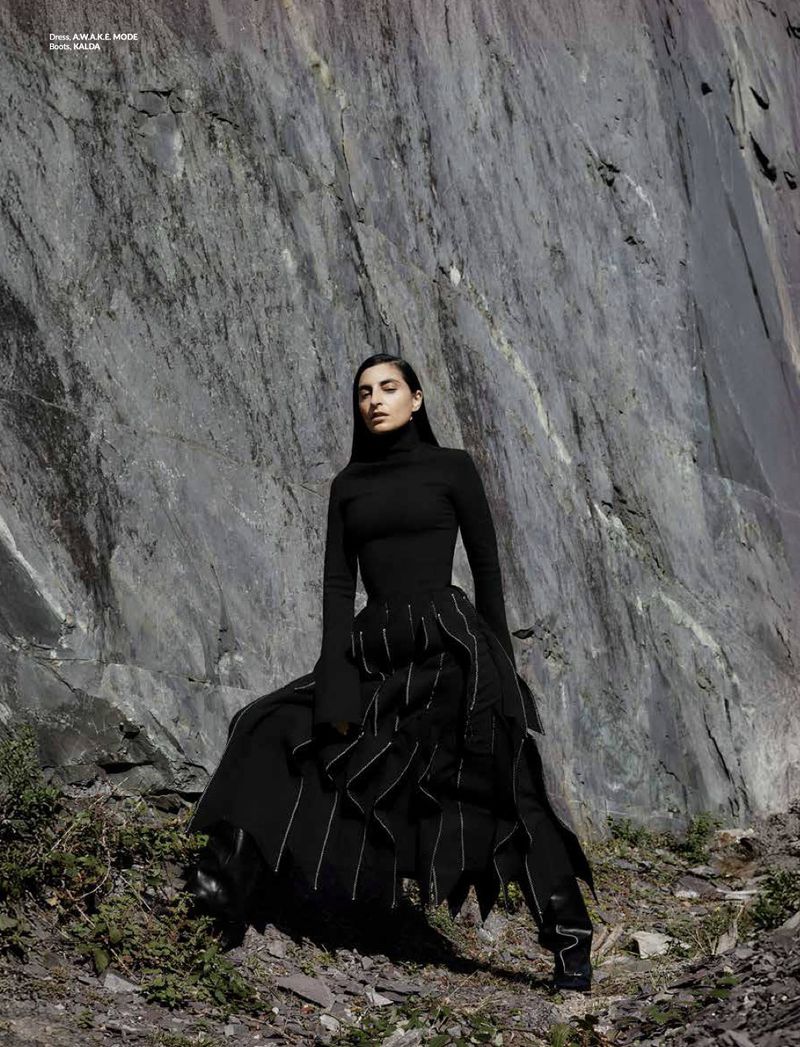 A lone female fashion model poses in a striking black outfit against a dramatic rocky mountain face during an outdoor fashion shoot.