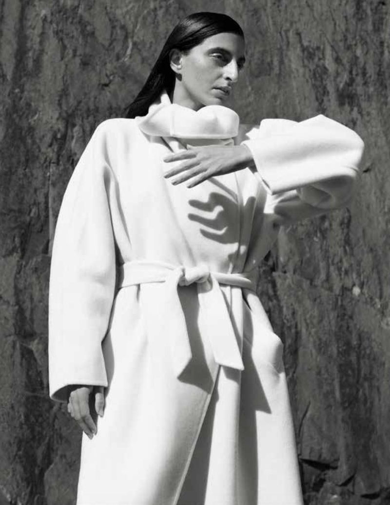 Model Nassia Matsa posing in an oversized white belted wrap coat with a high collar against a rugged rock wall.