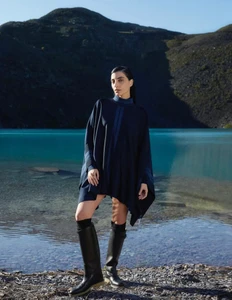Model in dark blue poncho and black tall boots standing beside a turquoise lake against a mountain backdrop.