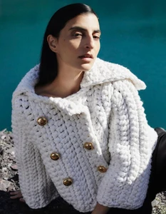 Model Nassia Matsa wearing a chunky white knit jacket with gold buttons against a vivid turquoise water background.