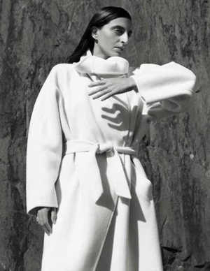 Model Nassia Matsa posing in an oversized white belted wrap coat with a high collar against a rugged rock wall.