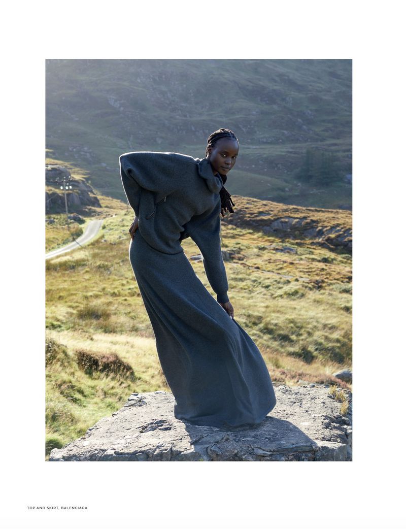 Fashion editorial shot of an African woman wearing a black Balenciaga dress, posing on a rock with mountains and a road in the background.