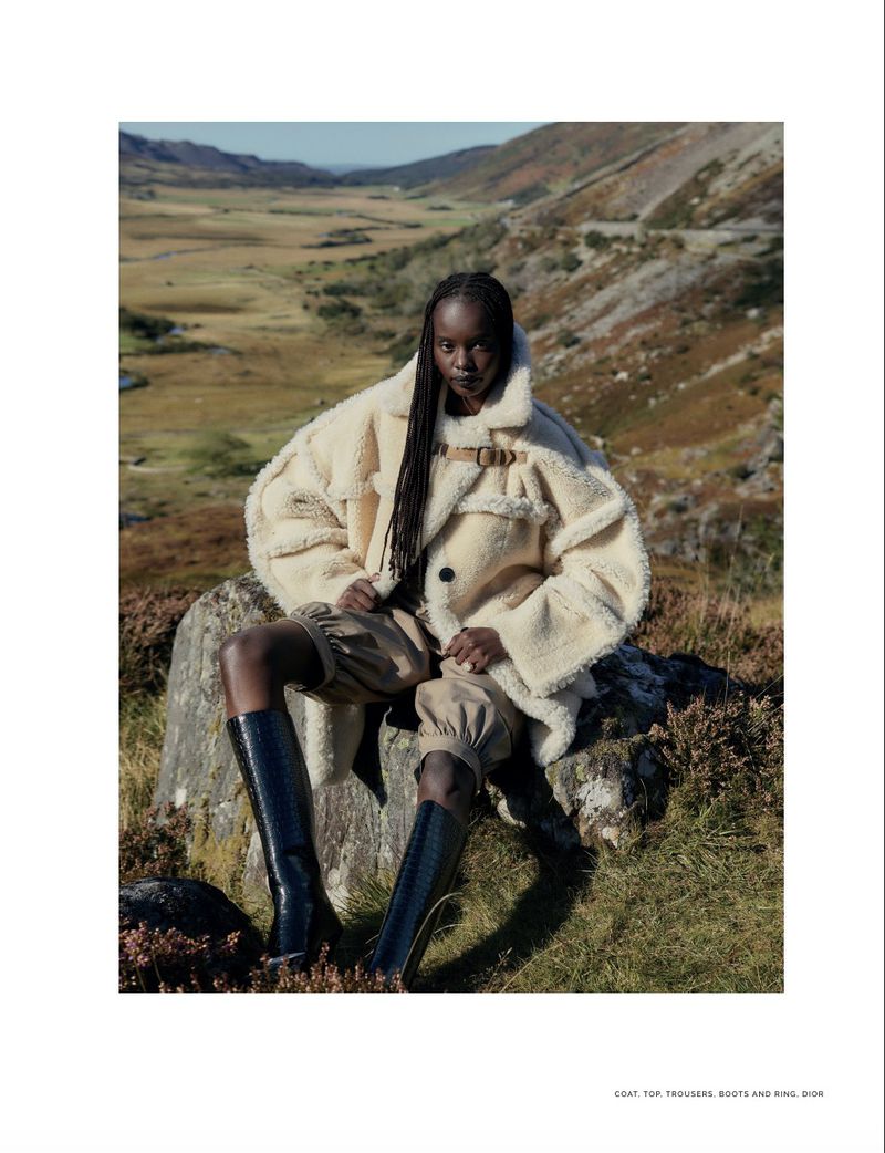 A black woman wearing a cream shearling coat and boots poses on a rock in a scenic mountain landscape. Fashion editorial shot outdoors.