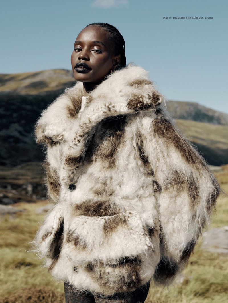 Fashion editorial portrait of a woman wearing a shaggy fur coat in a grassy field with mountains beneath a blue sky.