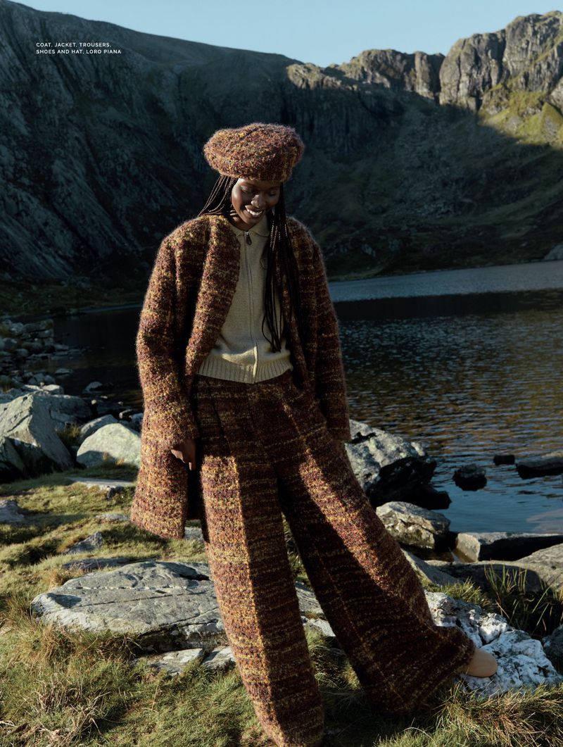 A smiling Black woman poses in a brown tweed suit and beret on a rocky lakeside with mountains in the background on a sunny day.