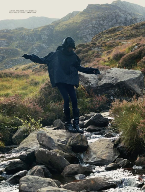 Woman in hooded poncho crossing rocky stream in a mountain landscape wearing dark trousers and boots in an outdoor fashion editorial.