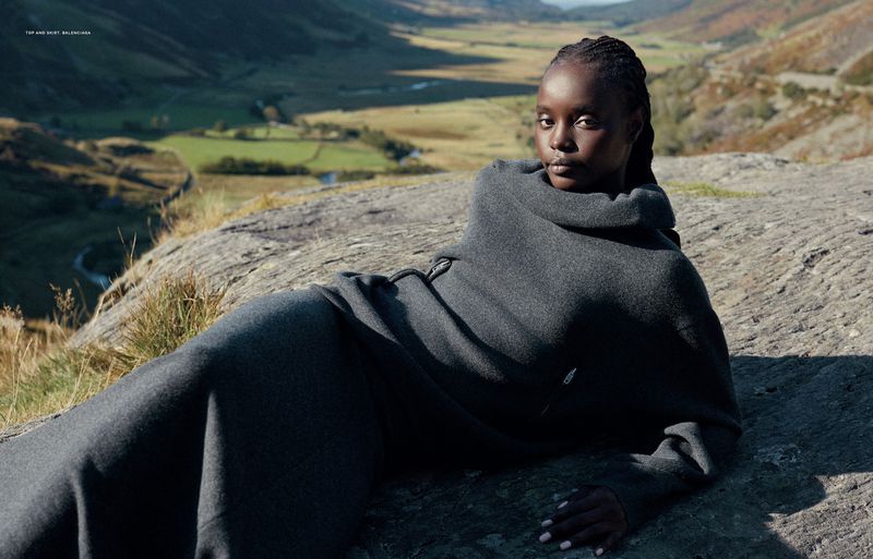 Woman lies on a rock in a gray outfit against a valley backdrop. Braided hair and direct gaze. Fashion editorial with natural outdoor setting.