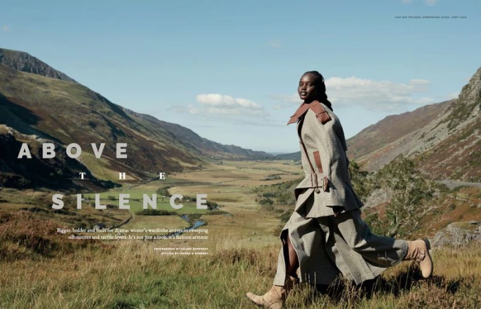 Fashion editorial shot of a Black female model wearing a houndstooth coat and skirt walking in a field against a mountain backdrop on a sunny day.