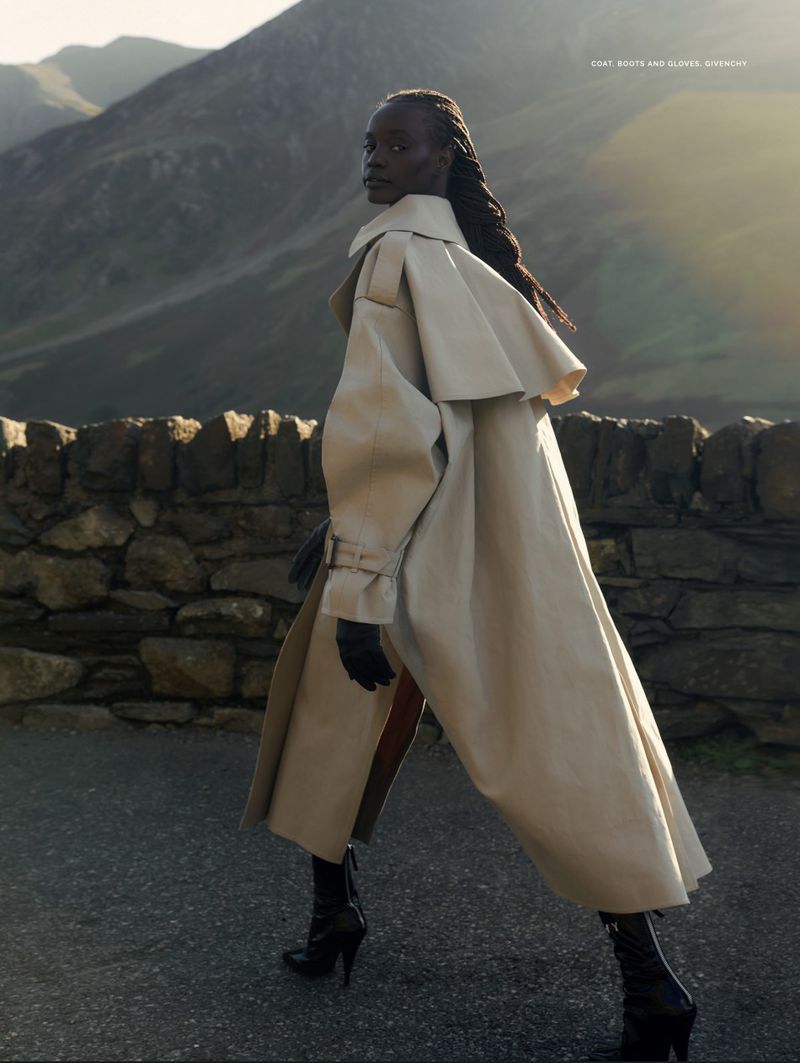 A woman in a beige coat walks on a road with mountains in the background, showcasing a blend of fashion and natural scenery.