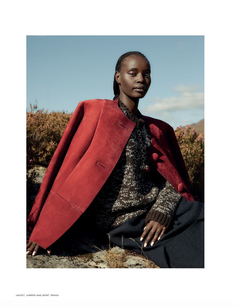 Fashion editorial shot of a Black woman in a red jacket and printed sweater sitting on rocks with a clear blue sky background.