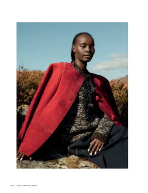 Model Achan Biong seated outdoors wearing a structured red jacket over a patterned knit sweater against a clear blue sky.