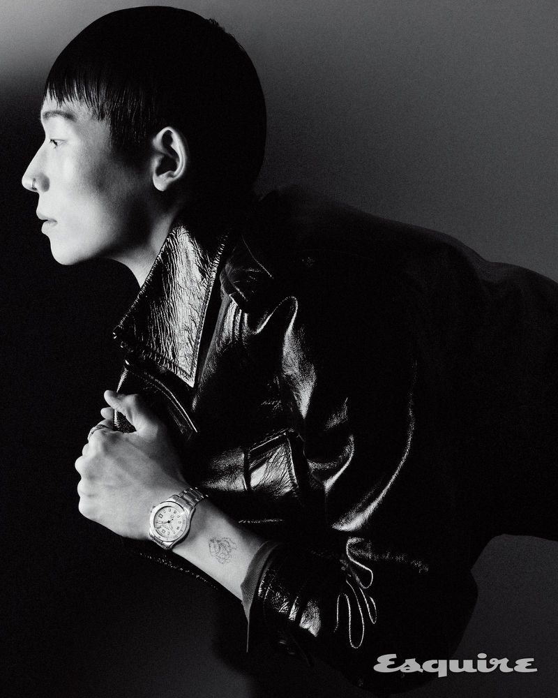 Monochrome portrait of a stylish adult man in a black leather jacket, posing with a watch on his wrist in a dark studio.