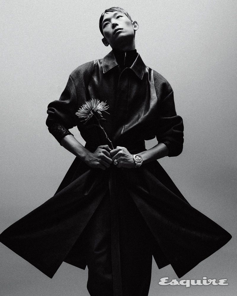 Asian male model in a black overcoat holds a flower, posed in a monochrome studio shot. The Esquire logo is visible at the bottom.