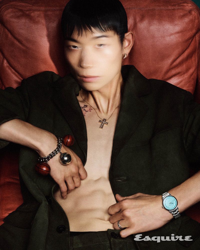 Portrait of a young Asian man posing on a red leather chair, wearing an open black jacket, jewelry, and a watch in an indoor setting.