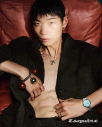 Close-up portrait of a male model wearing a dark jacket, wristwatch, and jewelry against a red leather armchair