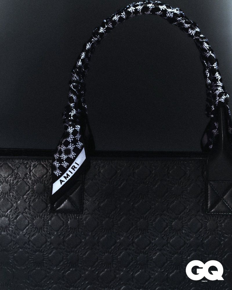 Close-up of a black textured leather tote bag with a patterned handle scarf, and a GQ magazine logo in the lower right corner.