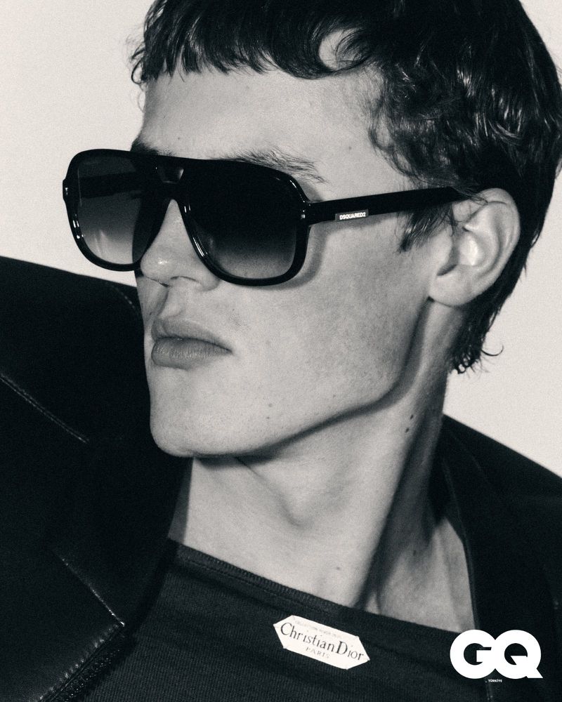 Monochromatic portrait of a man with dark hair wearing sunglasses and a black jacket, showcasing a Christian Dior label. GQ logo visible.