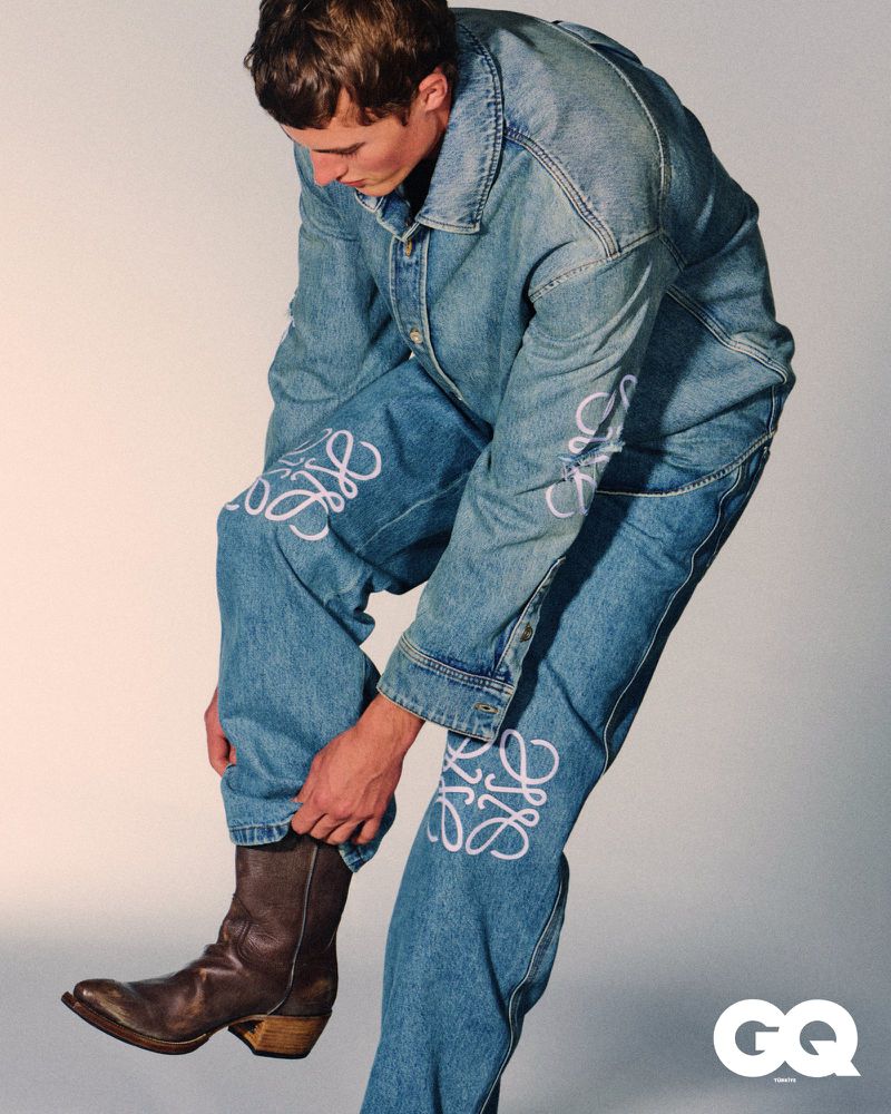 Fashion shot of a male model wearing a denim jacket and jeans with white Loewe branding, paired with brown cowboy boots, against a grey backdrop.