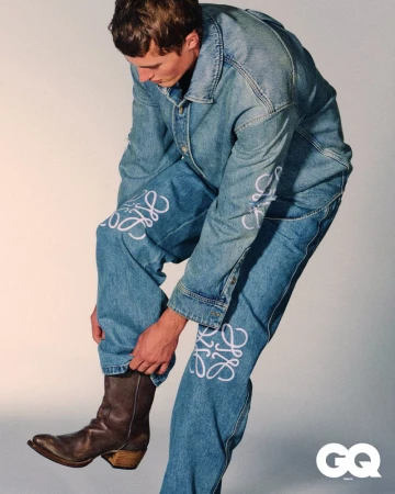 Fashion shot of a male model wearing a denim jacket and jeans with white Loewe branding, paired with brown cowboy boots, against a grey backdrop.