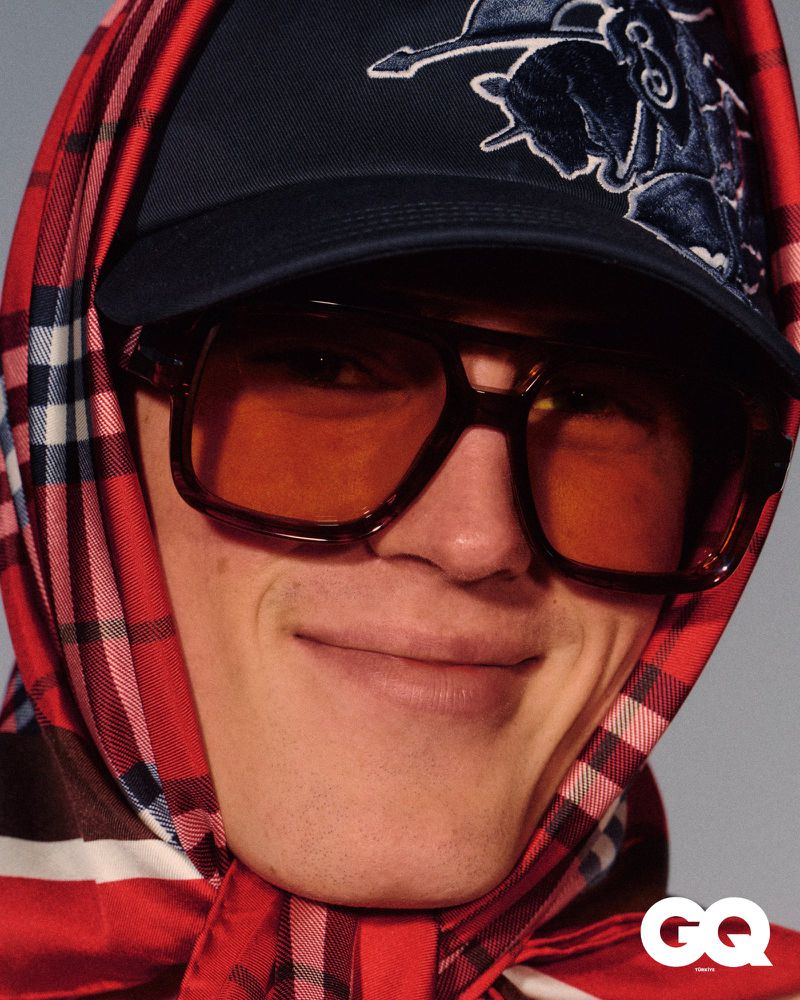 Portrait of a smiling adult male wearing a baseball cap, scarf, and sunglasses, close-up studio shot, fashion editorial.