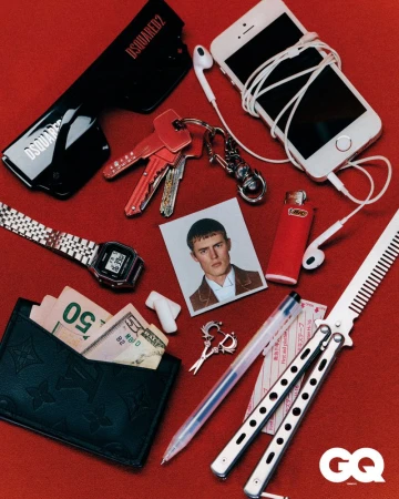 High-angle product shot on red background: black wallet with cash, smartphone, earphones, watch, keys, sunglasses, wallet, lighter, comb, pen.