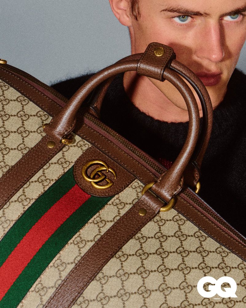 A stylish male model showcases a Gucci handbag with the brand's logo, set against a plain background in a fashion editorial photograph.