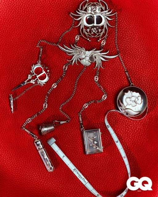 Jewellery set, including silver pendant necklaces and a tape measure on red leather. Logo present in the corner.