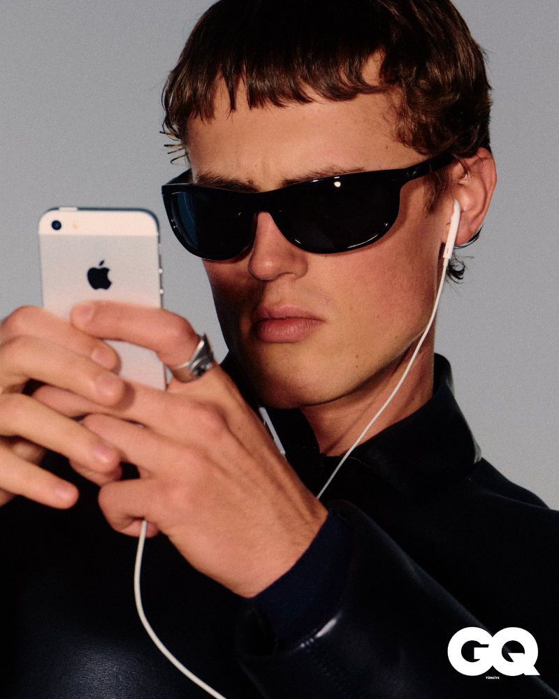 Portrait of a young adult male wearing sunglasses, wired headphones, and holding an iPhone in a studio setting with simple lighting.