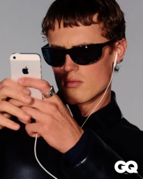Young man wearing dark sunglasses and black jacket looking at a white smartphone with wired earphones in a studio shoot.