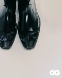 High-angle close up of black glossy square-toe boots with knotted satin details across the toe on a pale neutral backdrop.