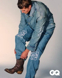 Young man wearing a denim shirt and printed jeans pulling on a brown leather boot against a light grey background