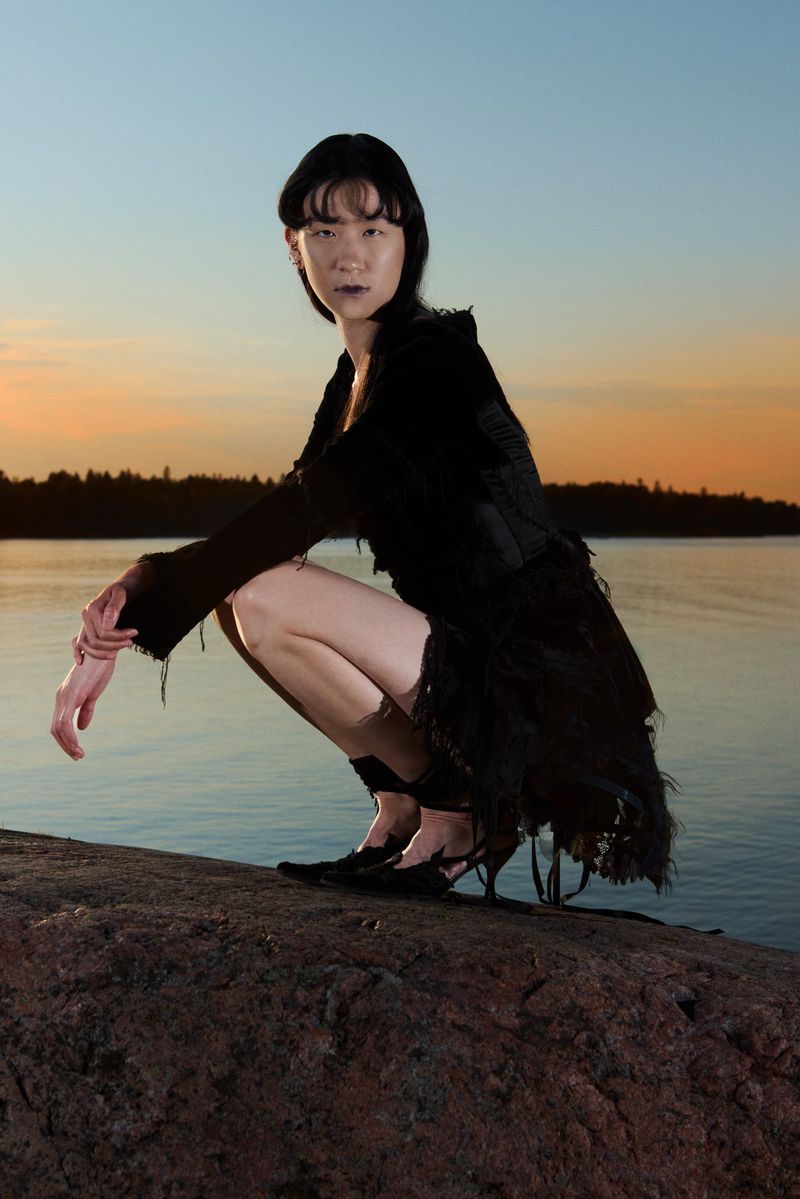 Model crouching on rocky shoreline wearing a black dress against a sunset sky and water background