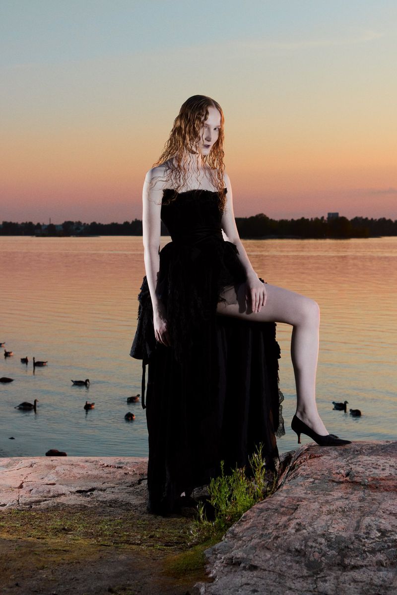A woman in a black dress stands on rocks near a lake at sunset, fashion photography, dark and elegant style, water reflection, outdoor location.