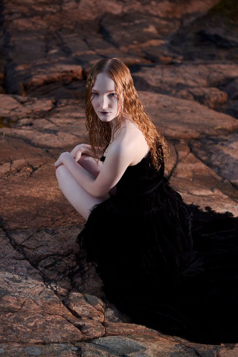 A young woman with red hair is posed crouching on rocks wearing a long black gown in a fashion portrait taken outdoors.