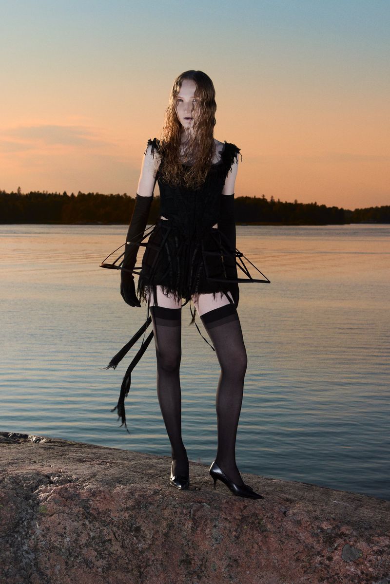 A slender model stands on a rock by a calm lake at sunset, wearing a black outfit with gloves, stockings, and an unusual frame design.
