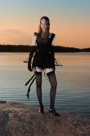A slender model stands on a rock by a calm lake at sunset, wearing a black outfit with gloves, stockings, and an unusual frame design.