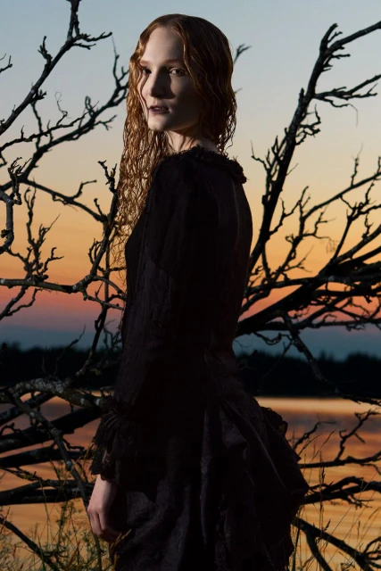 Red-haired woman in a vintage dress, standing outdoors among bare branches at dusk. A dramatic, dark, and romantic visual.