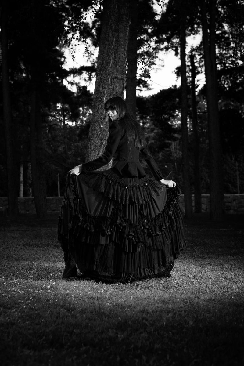 A woman in a dark Victorian-style dress stands among trees in a forest, holding the sides of her tiered, ruffled skirt in a vintage outdoor scene.