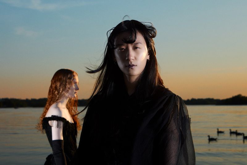 Two women in dark dresses stand on a tranquil lake at sunset. The water reflects the fading light, creating a peaceful, ethereal atmosphere.