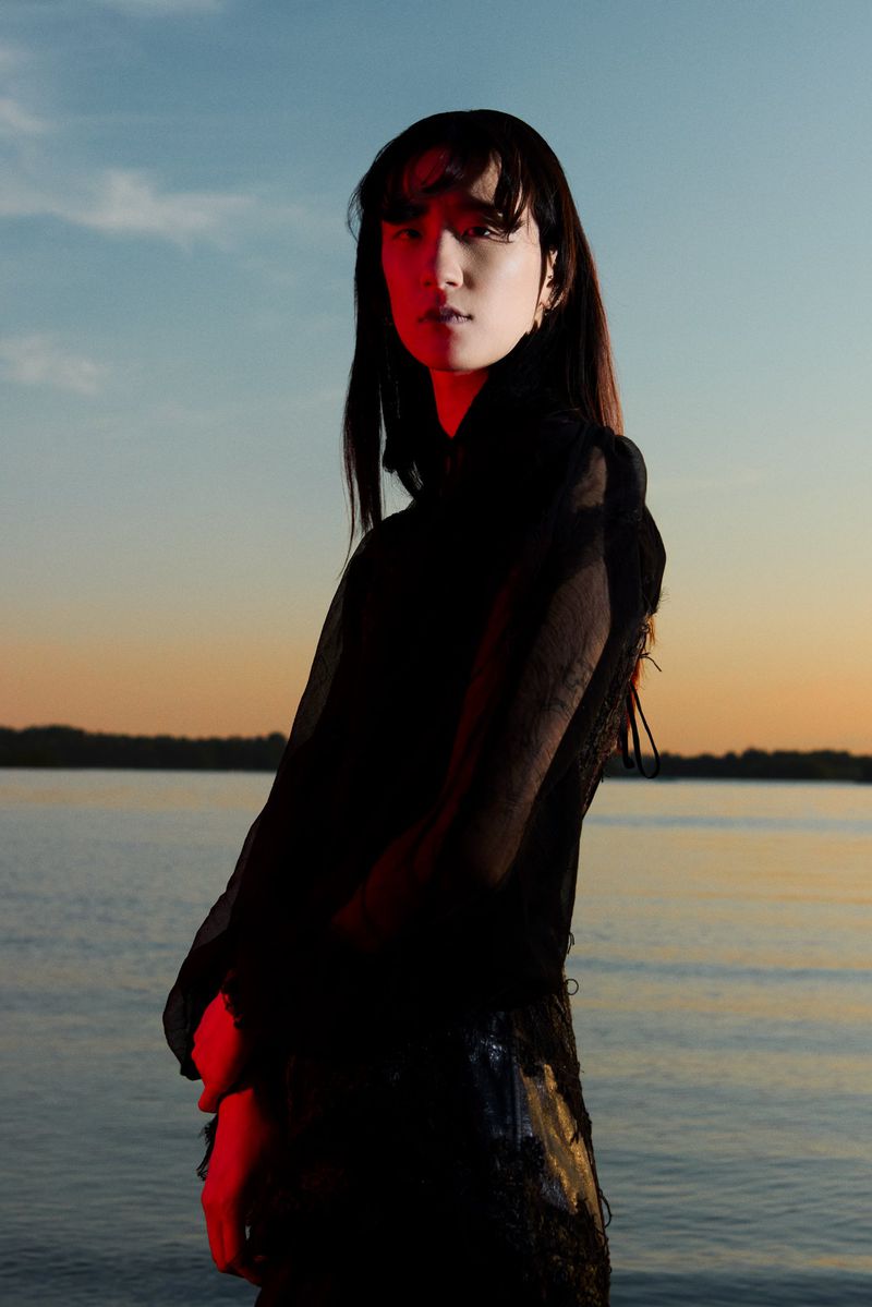 Model Yilin Ma standing in a sheer black top at dusk against calm water with red light cast on her face and hands.