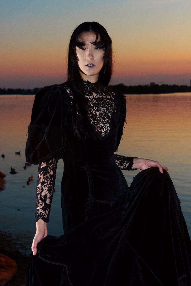 Portrait of a person in a black Victorian-style dress by a lake at sunset, creating a melancholic and elegant fashion image.