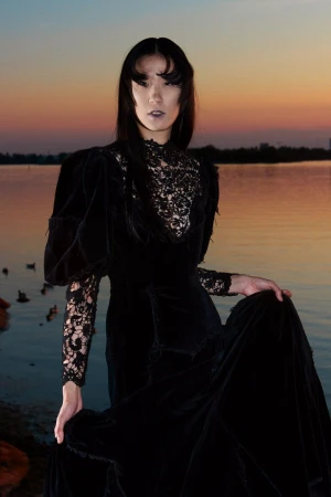 Portrait of a person in a black Victorian-style dress by a lake at sunset, creating a melancholic and elegant fashion image.