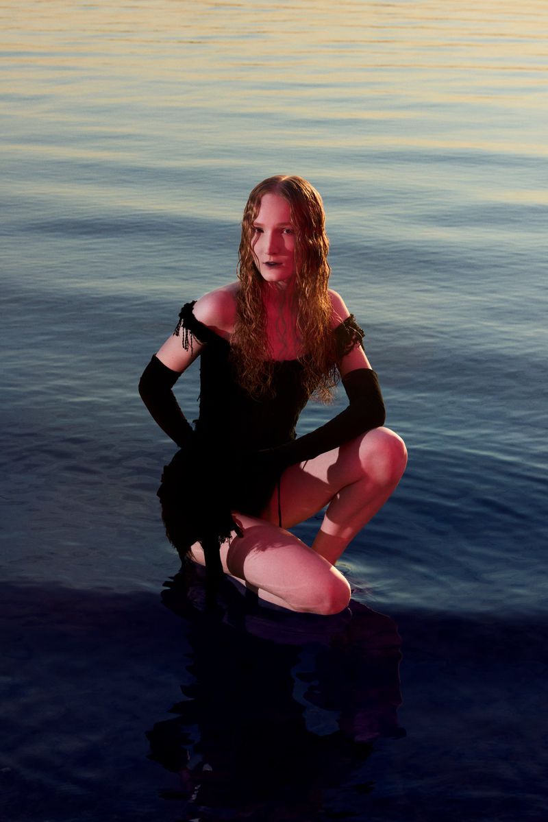 A model kneels in a black dress on a shore with water and sky visible. The model has long hair and poses facing the camera.