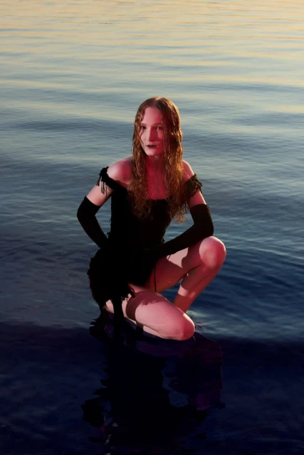 A model kneels in a black dress on a shore with water and sky visible. The model has long hair and poses facing the camera.