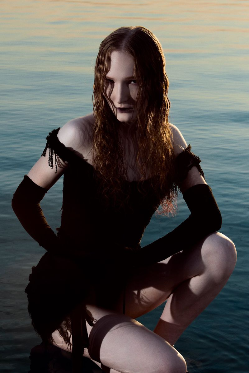 Woman in a black dress poses crouching against a blue ocean backdrop, featuring wet curly hair and striking dark lipstick.