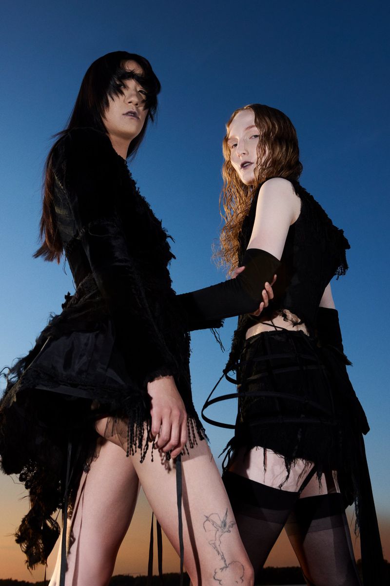 Two alternative fashion models in black gothic attire pose against a blue sky. The low angle emphasizes their dramatic style.
